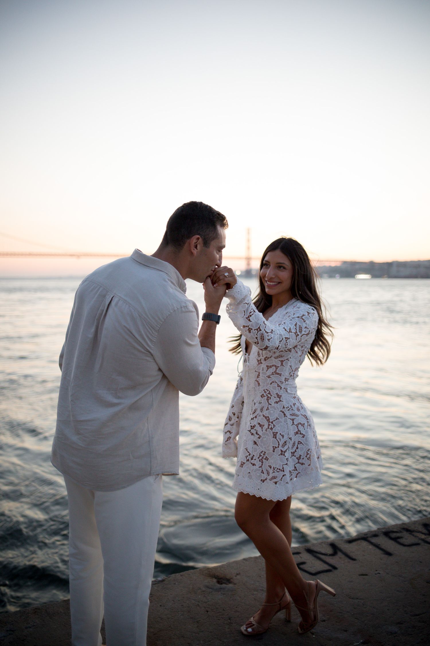 Man in white kisses woman's hand at sunset by the sea, both dressed in white, romantic beach proposal moment.
