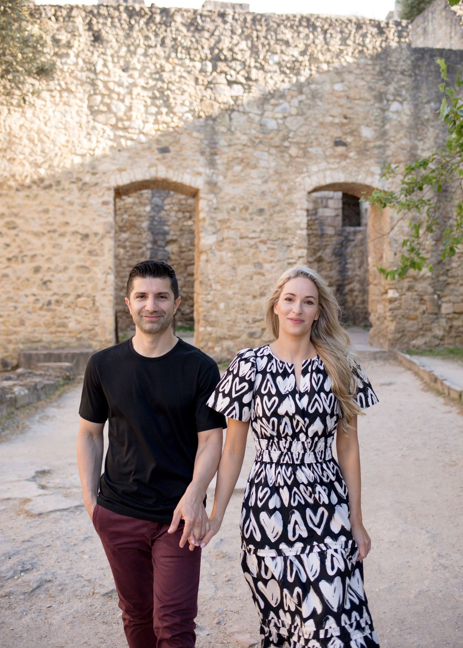 Couple holding hands walking near ancient stone ruins, woman in heart-print dress, man in black shirt and red pants.
