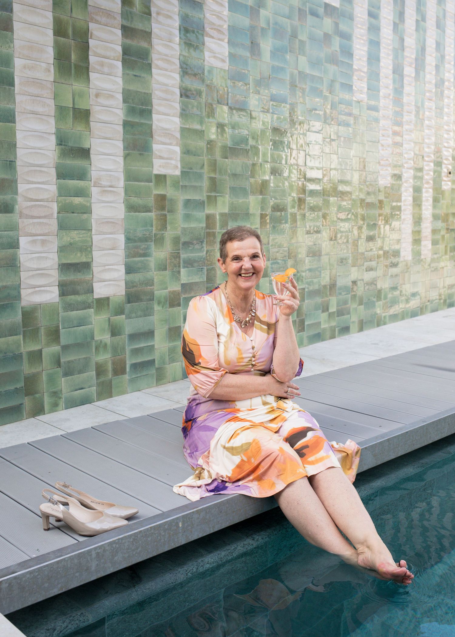 Smiling woman in a colorful floral dress sits at a pool's edge, feet in the water, holding an orange cocktail by a green tiled wall.