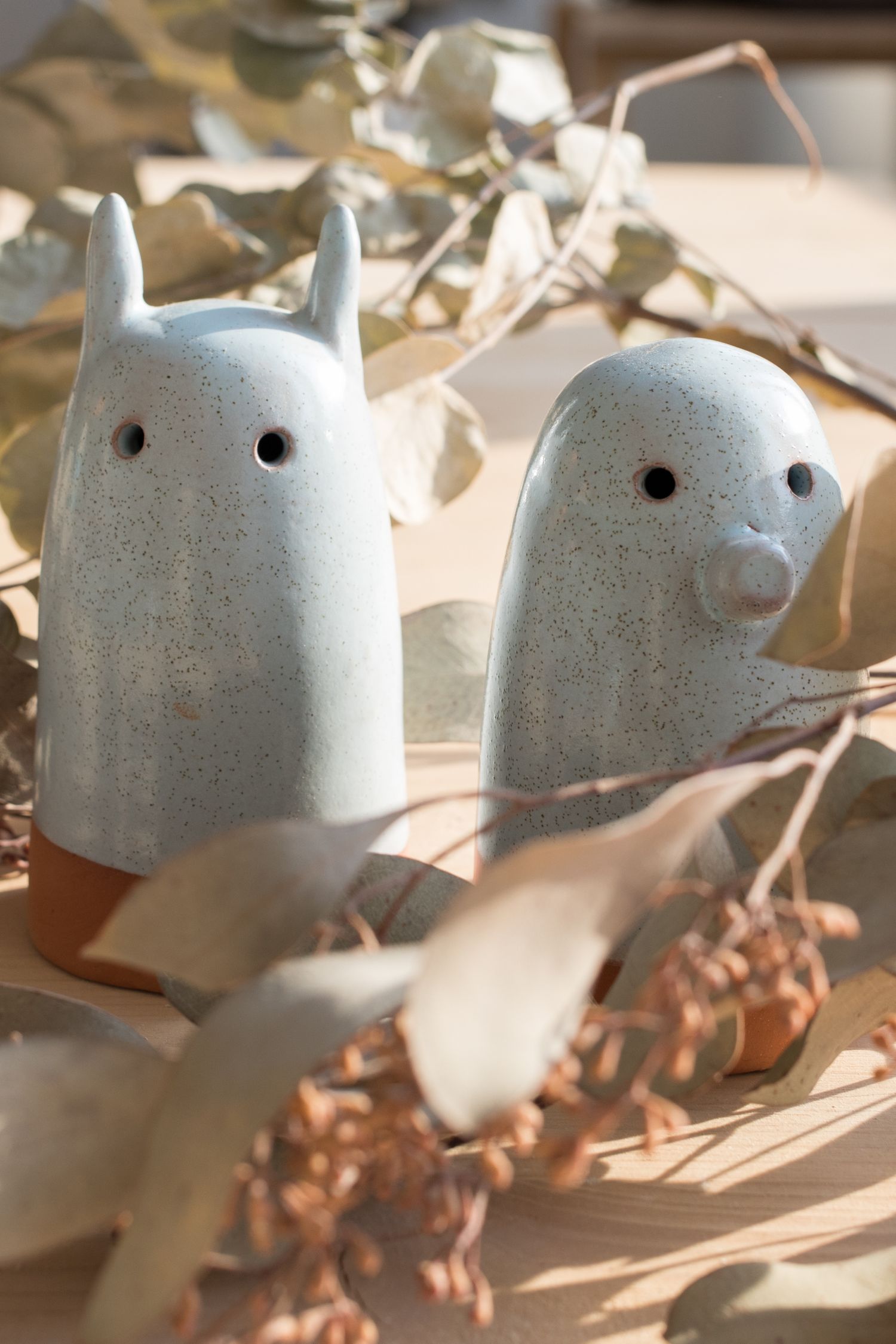 Two speckled white ceramic ghost figurines with terracotta bases, one with pointed ears and one with a round nose, among dried eucalyptus.