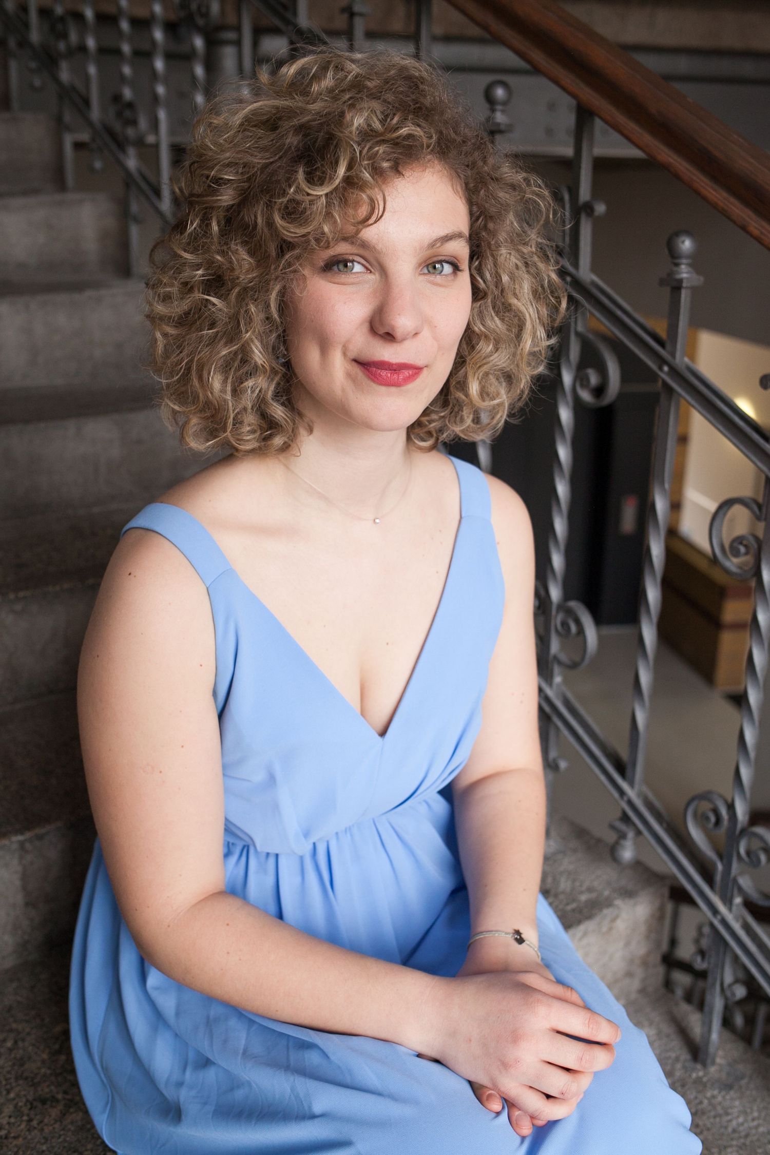 Smiling woman with curly hair in a light blue sleeveless dress sitting on indoor stairs beside an ornate wrought iron railing.