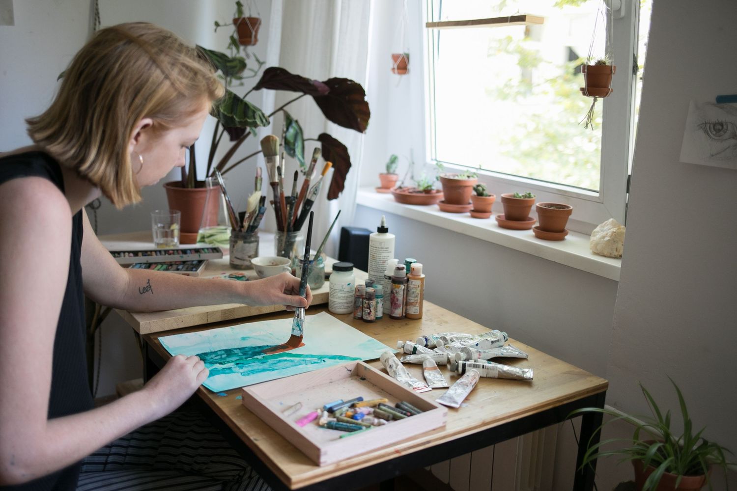 Woman painting with watercolors at a wooden desk by a sunny window, surrounded by brushes, paint supplies, and potted plants.