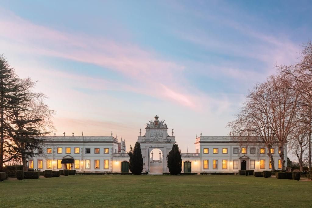 Grand neoclassical estate with illuminated wings, ornate central arch, and manicured lawn at dusk under a pink sky.