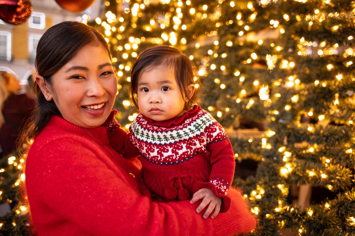A mother and baby in matching red Christmas sweaters smile in front of a glowing holiday tree.