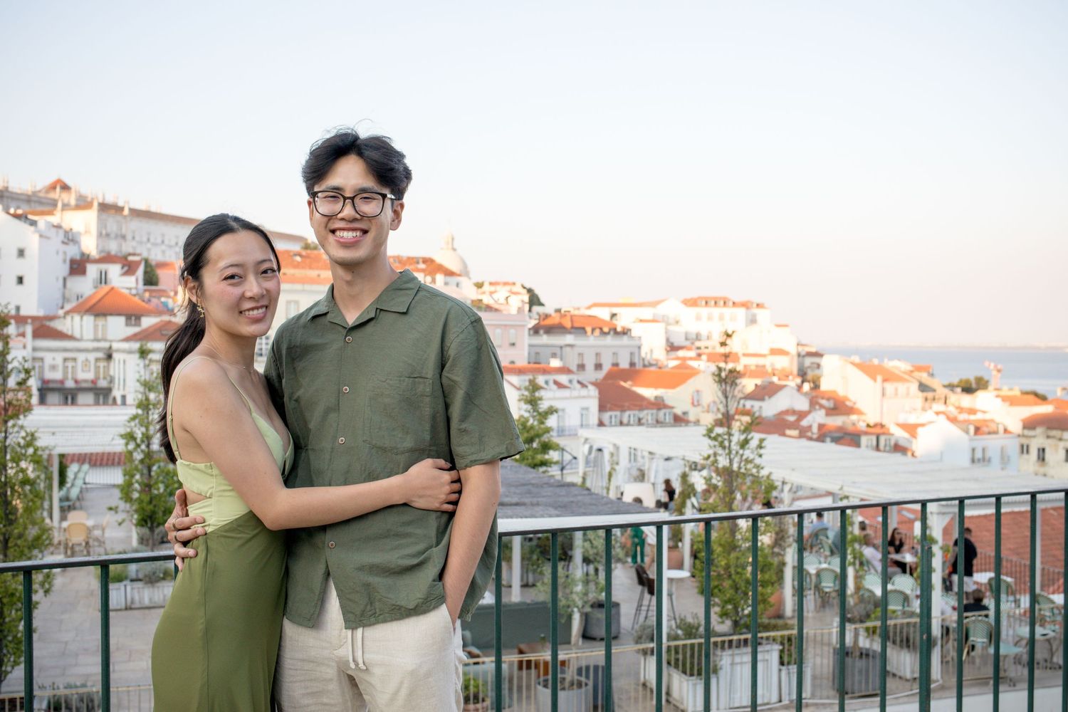 Couple smiling on a scenic rooftop terrace overlooking Lisbon's colorful cityscape at sunset.