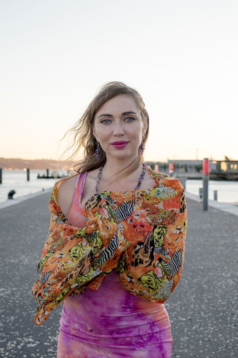Blonde woman in colorful floral wrap and pink dress posing on waterfront pier at sunset.