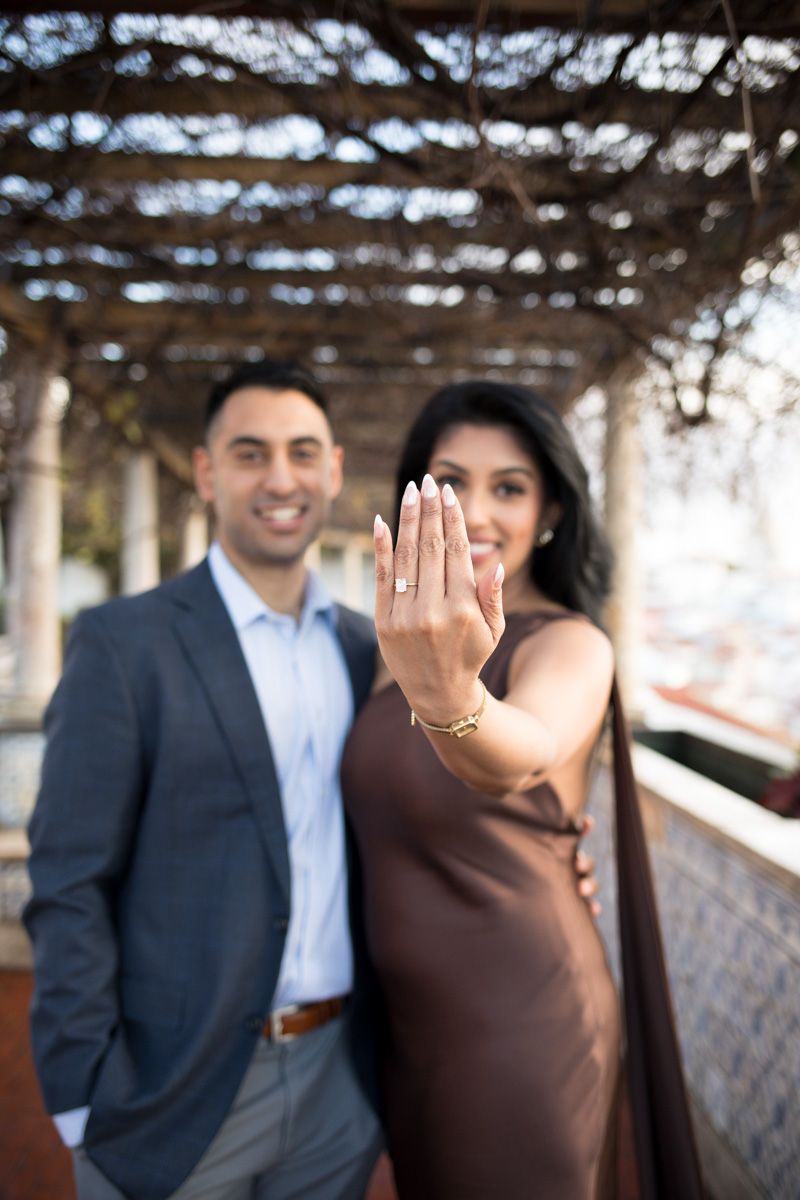 Couple showing off engagement ring under pergola, woman in brown dress extending hand with diamond ring toward camera.