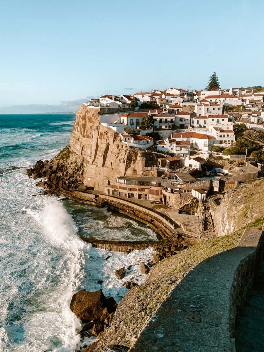 Coastal village perched on rocky cliffs above crashing Atlantic waves, white houses with red roofs under clear blue sky.