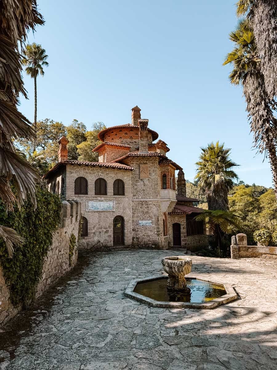 Stone villa with octagonal fountain courtyard, terracotta roof, arched windows, surrounded by palm trees under clear blue sky.