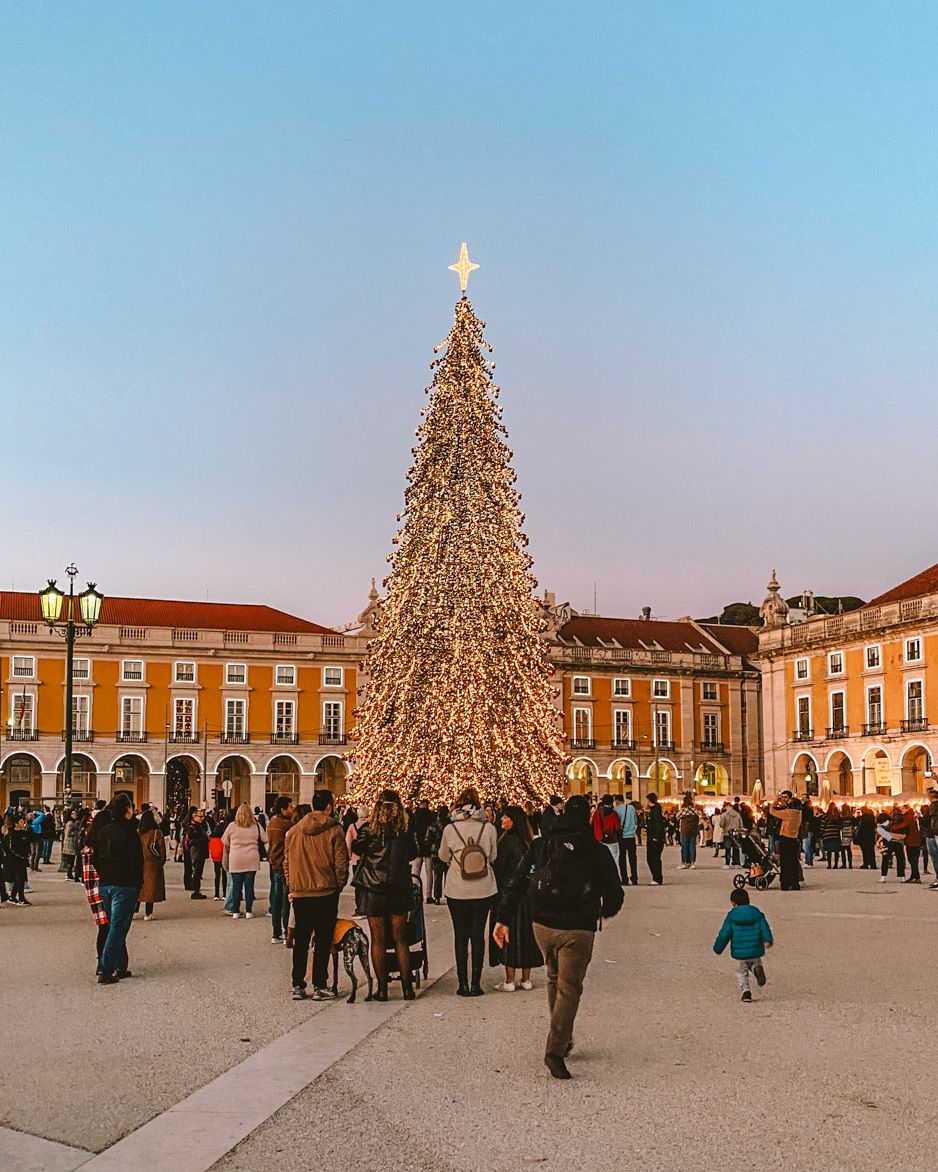 A large illuminated Christmas tree in Lisbon's Praca do Comercio square, surrounded by crowds at dusk.