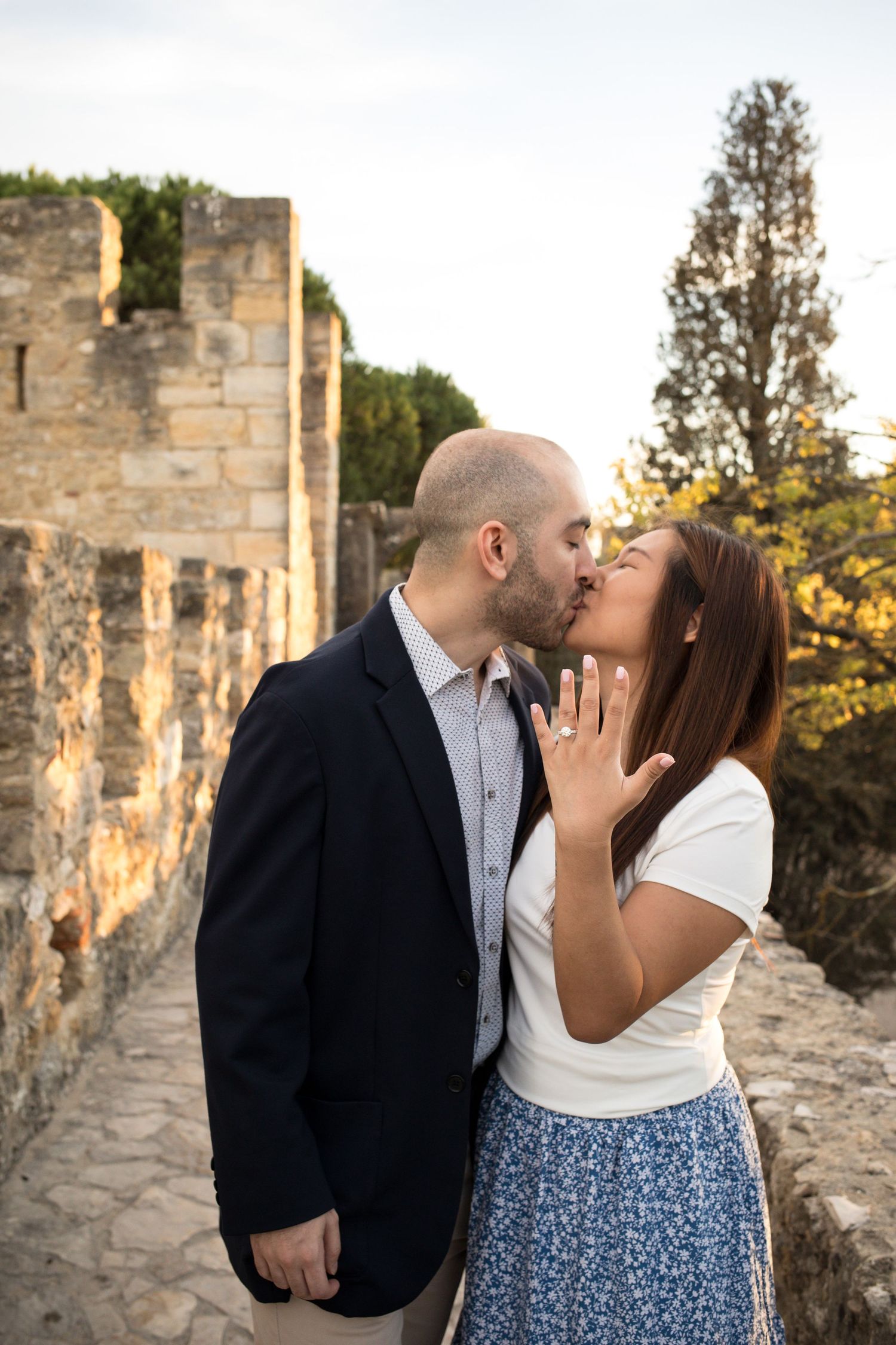Couple sharing a kiss during engagement, woman showing diamond ring, standing on historic stone castle walls.