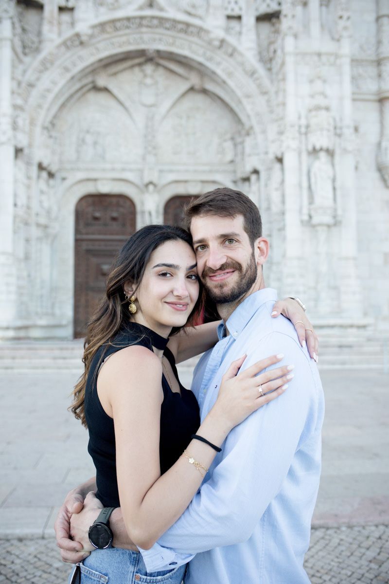 A smiling couple embracing in front of an ornate Gothic cathedral facade, woman in black top with gold earrings.