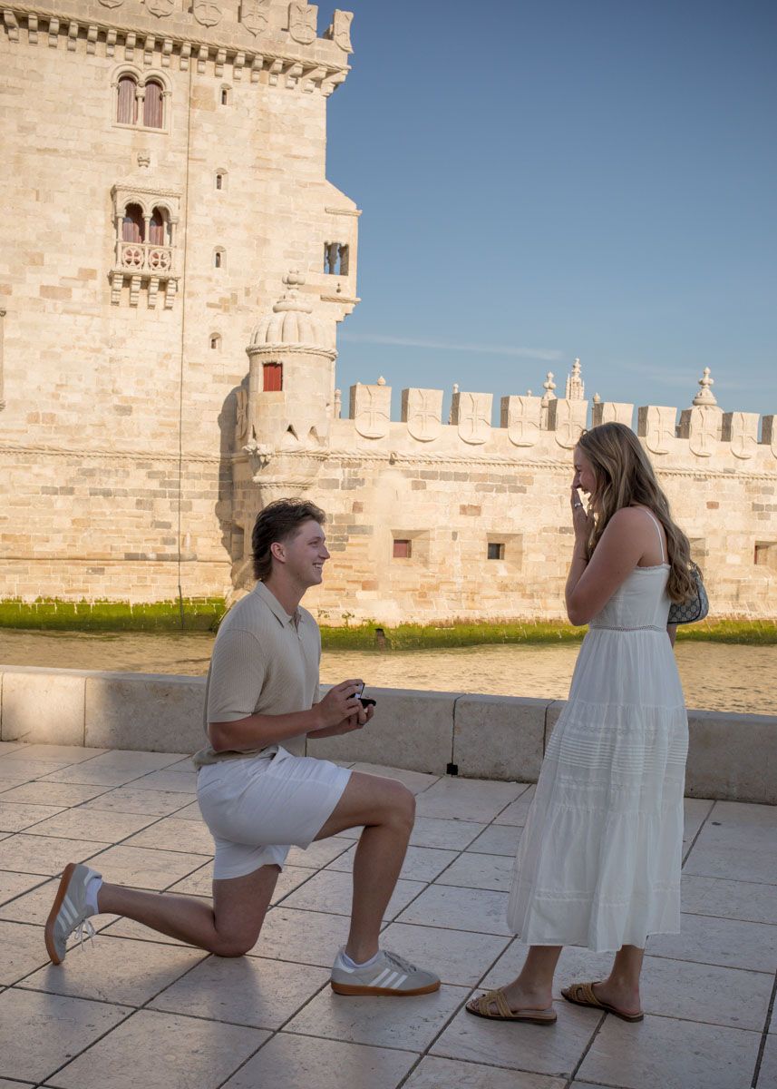 Man proposes on one knee to woman in white dress at Belem Tower, Lisbon, Portugal at golden hour.