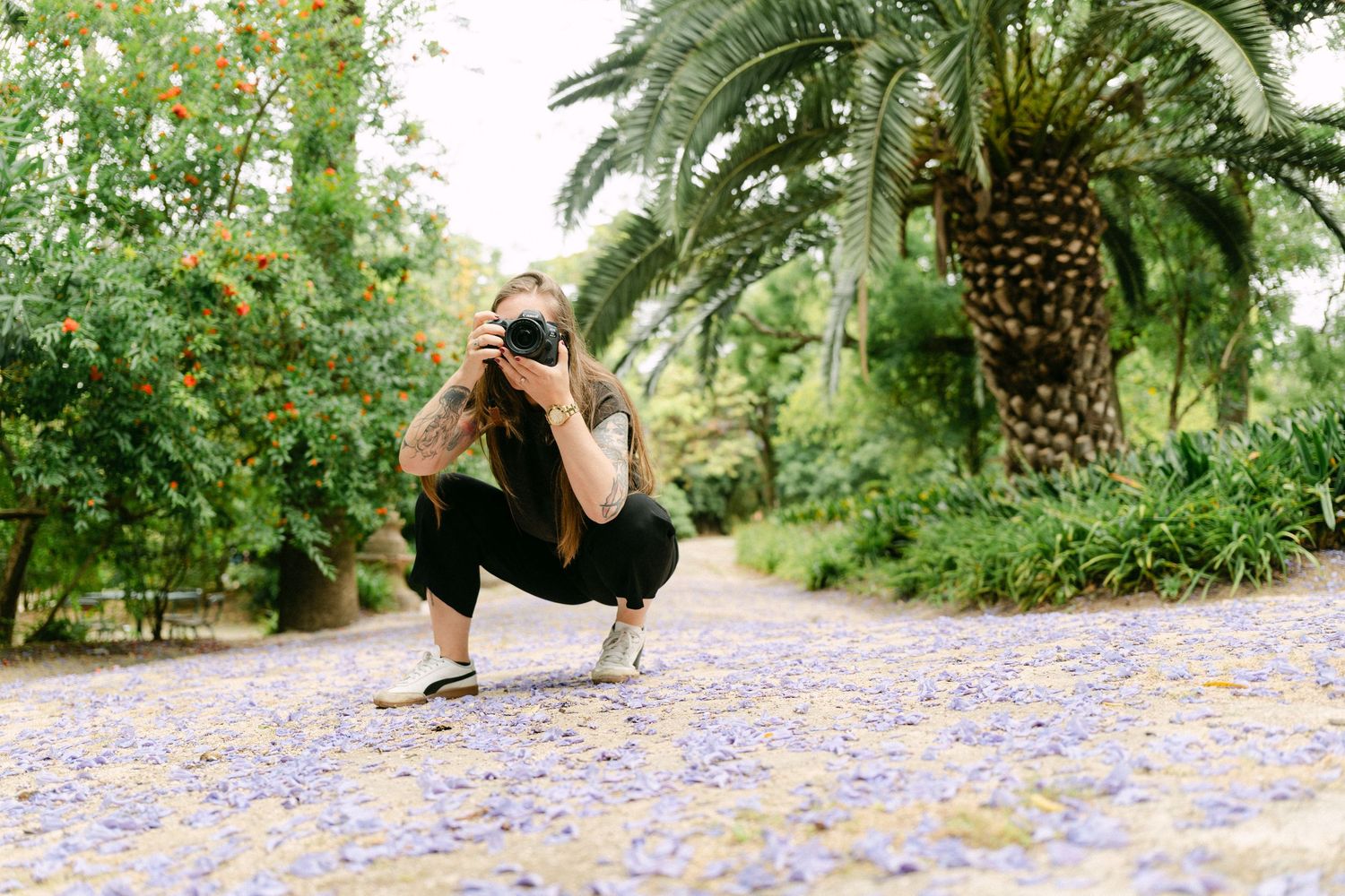 Lisbon Photographer crouching on purple flower-covered path in lush garden with palm trees, capturing photos with DSLR camera.