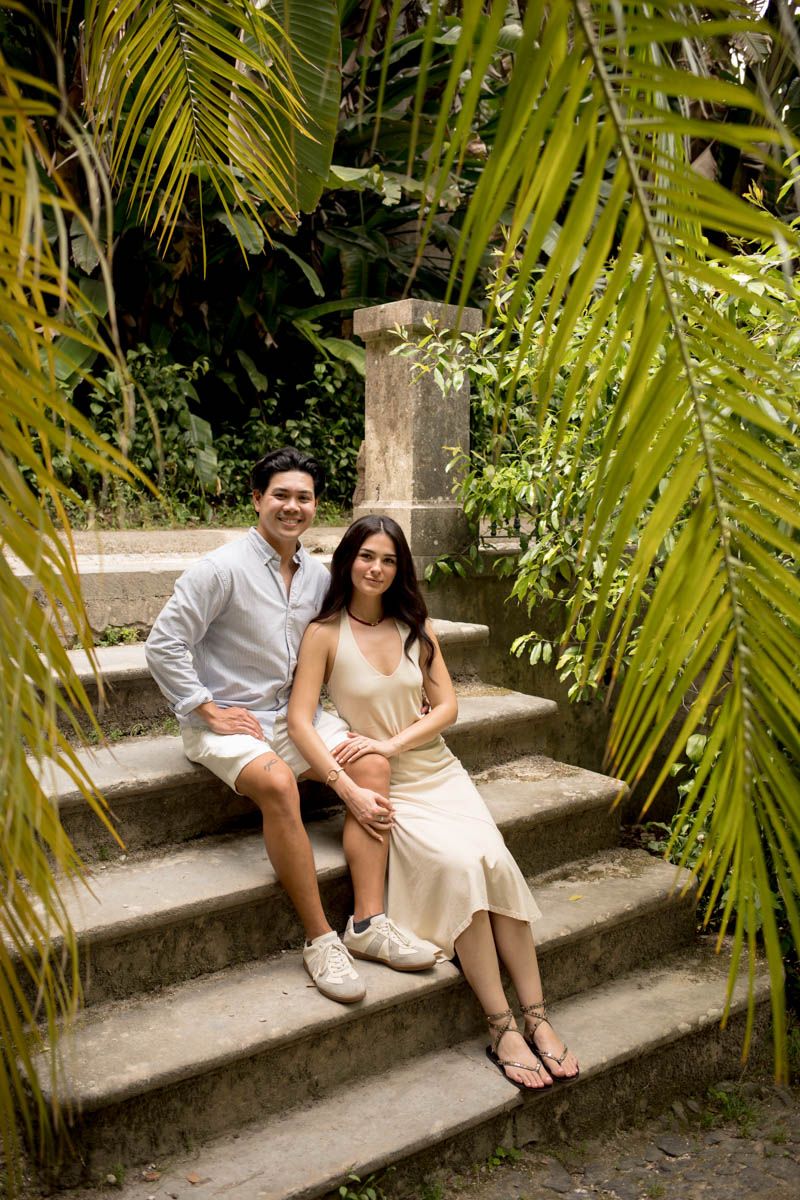 Smiling couple sits together on stone steps surrounded by lush tropical palm fronds in a garden setting.