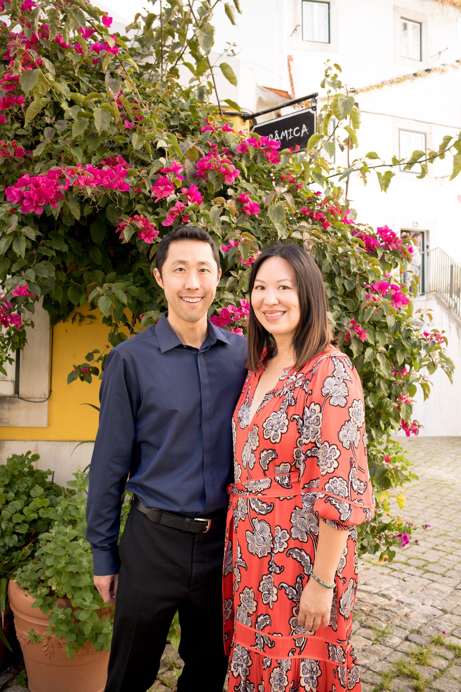 Smiling couple standing together in front of vibrant pink bougainvillea flowers at a charming European street.