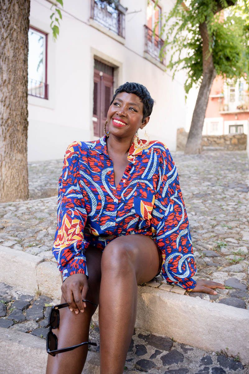 Happy woman in vibrant printed blouse sitting on stone steps on cobblestone street.