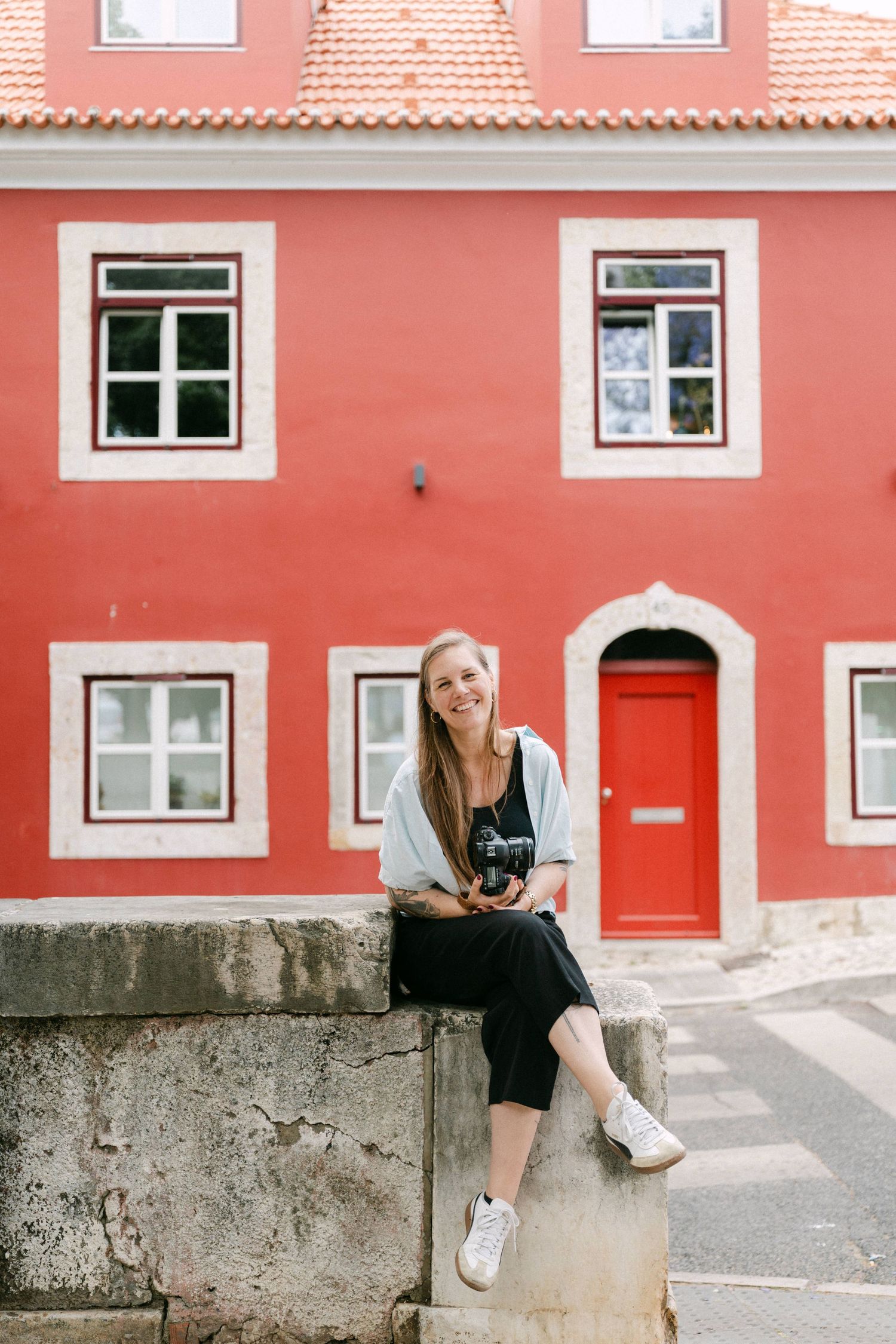 Smiling female photographer in Lisbon sitting on a stone wall holding a camera in front of a vibrant red building with white windows.