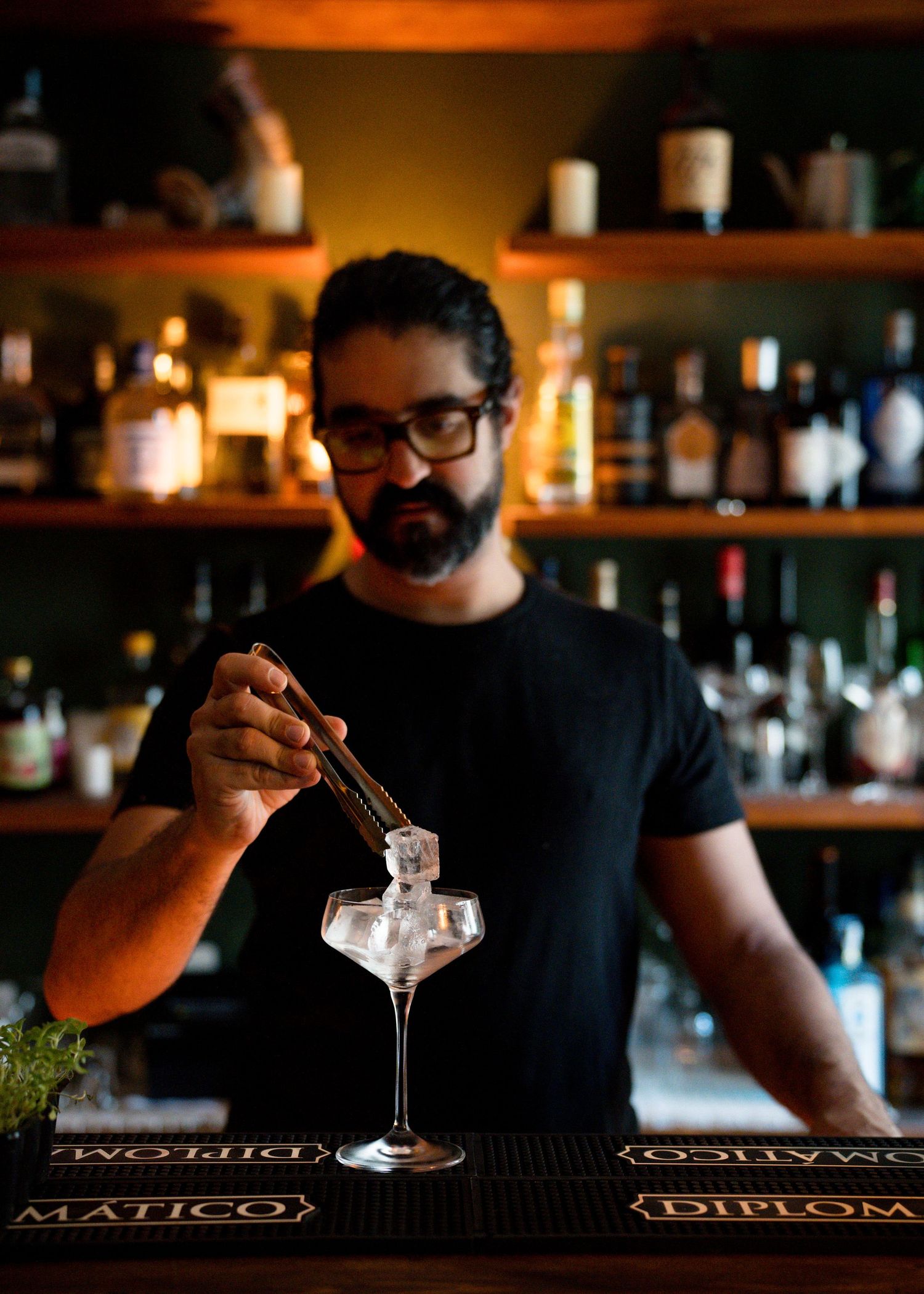 Bearded bartender in glasses uses tongs to place ice into a coupe glass at a bar with shelves of liquor bottles behind him.