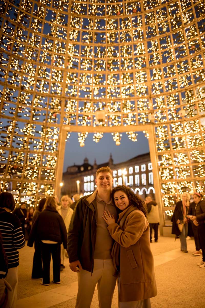 Couple smiling under a glowing dome of fairy lights at a festive outdoor Christmas market at night.