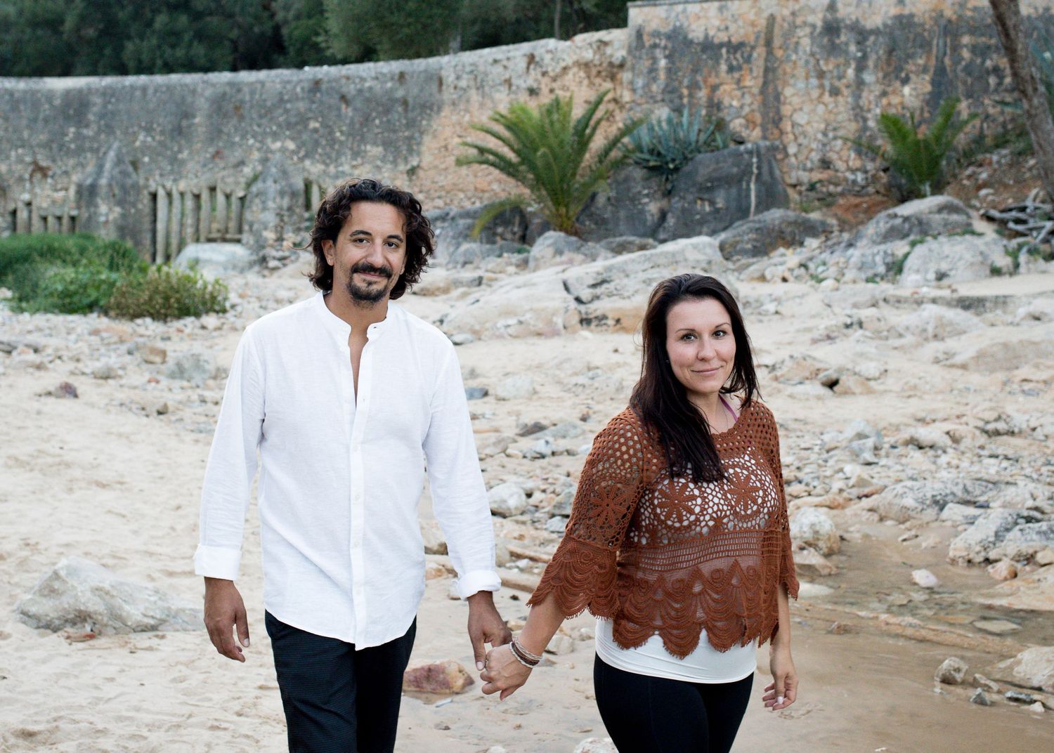 Couple walks hand in hand along a sandy beach with ancient stone ruins and palm trees in background.