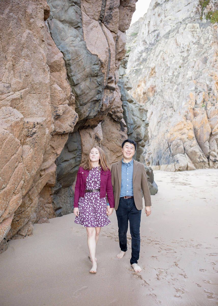 Happy couple walks barefoot on sandy beach between towering dramatic rock formations.