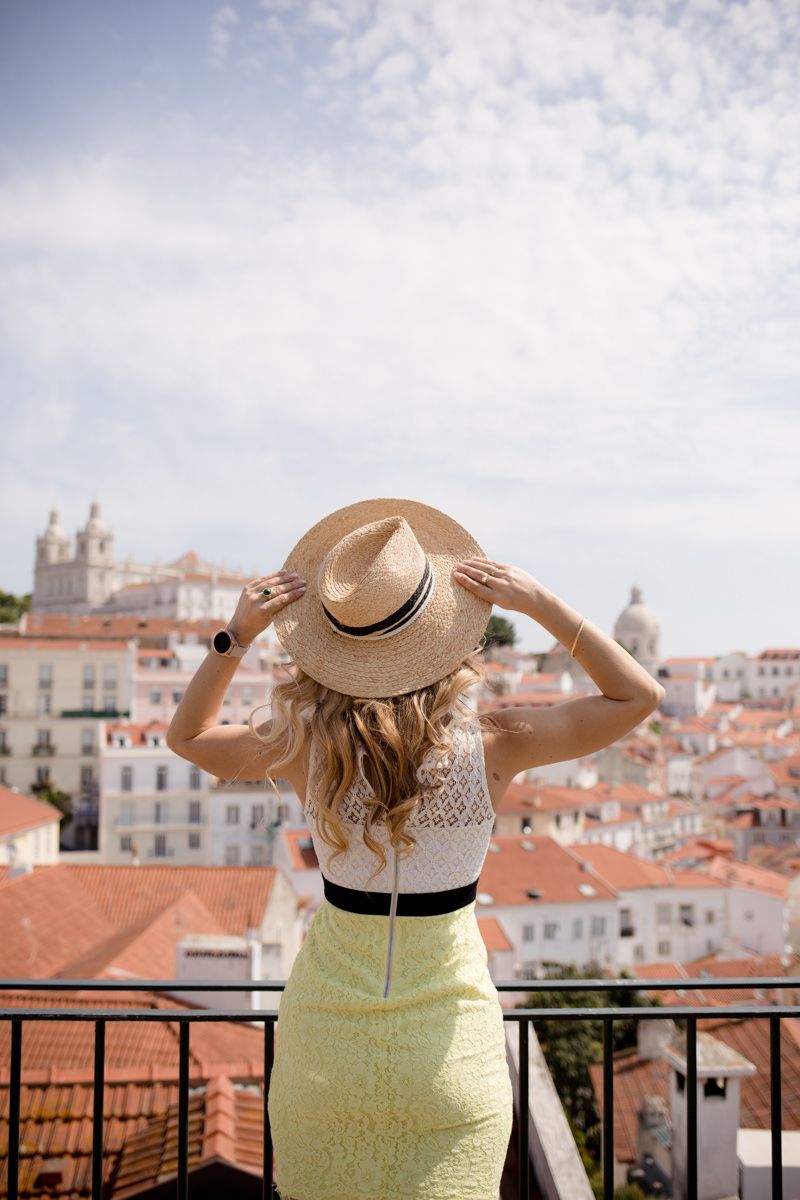 Blonde woman in straw hat and yellow skirt overlooks Lisbon rooftops from scenic viewpoint balcony.