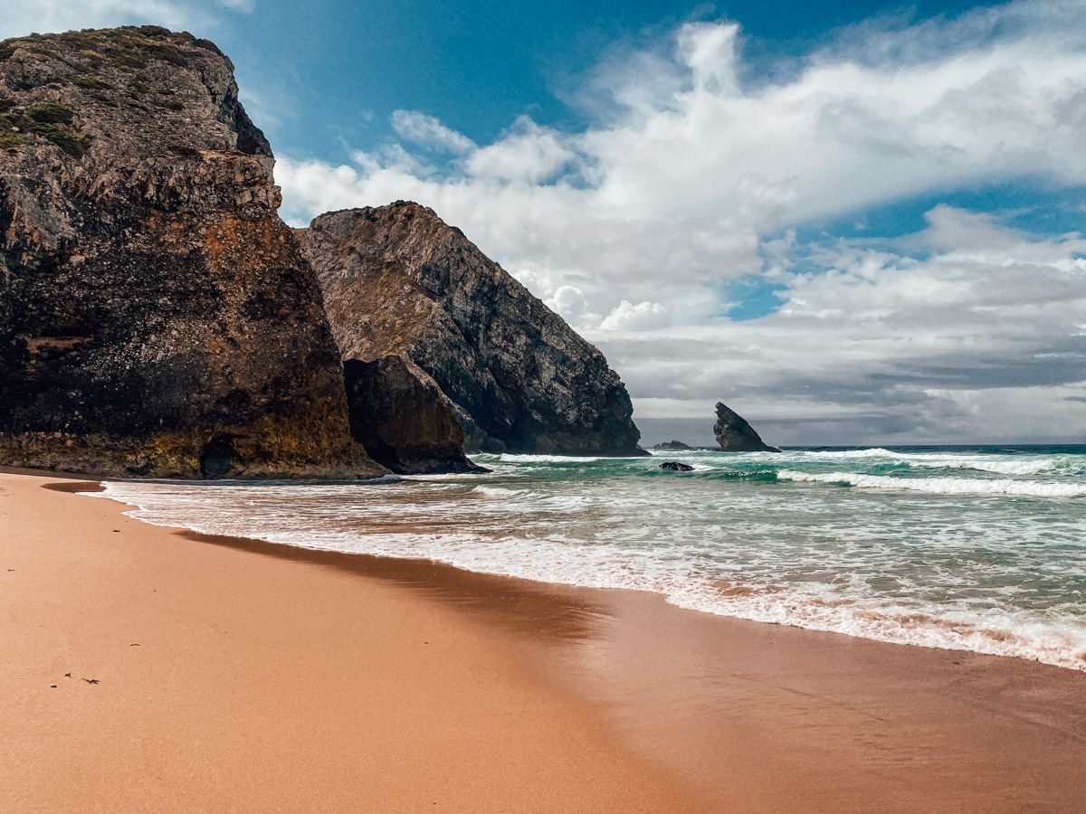 Sandy beach with dramatic rocky cliffs, ocean waves, and a partly cloudy sky on a scenic coastline.
