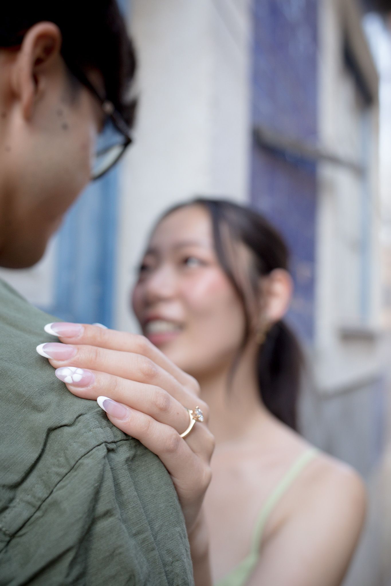 Smiling Asian woman places her hand on a man's shoulder, showcasing a gold ring with French manicured nails.