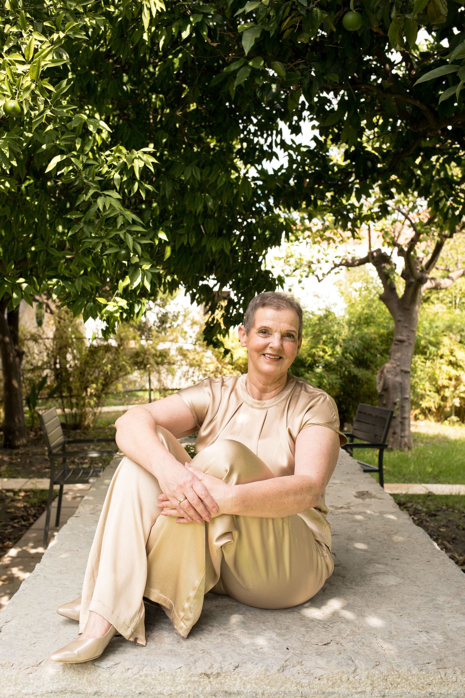 Smiling woman in a champagne satin outfit sits on a stone ledge in a sunlit garden beneath leafy citrus trees.