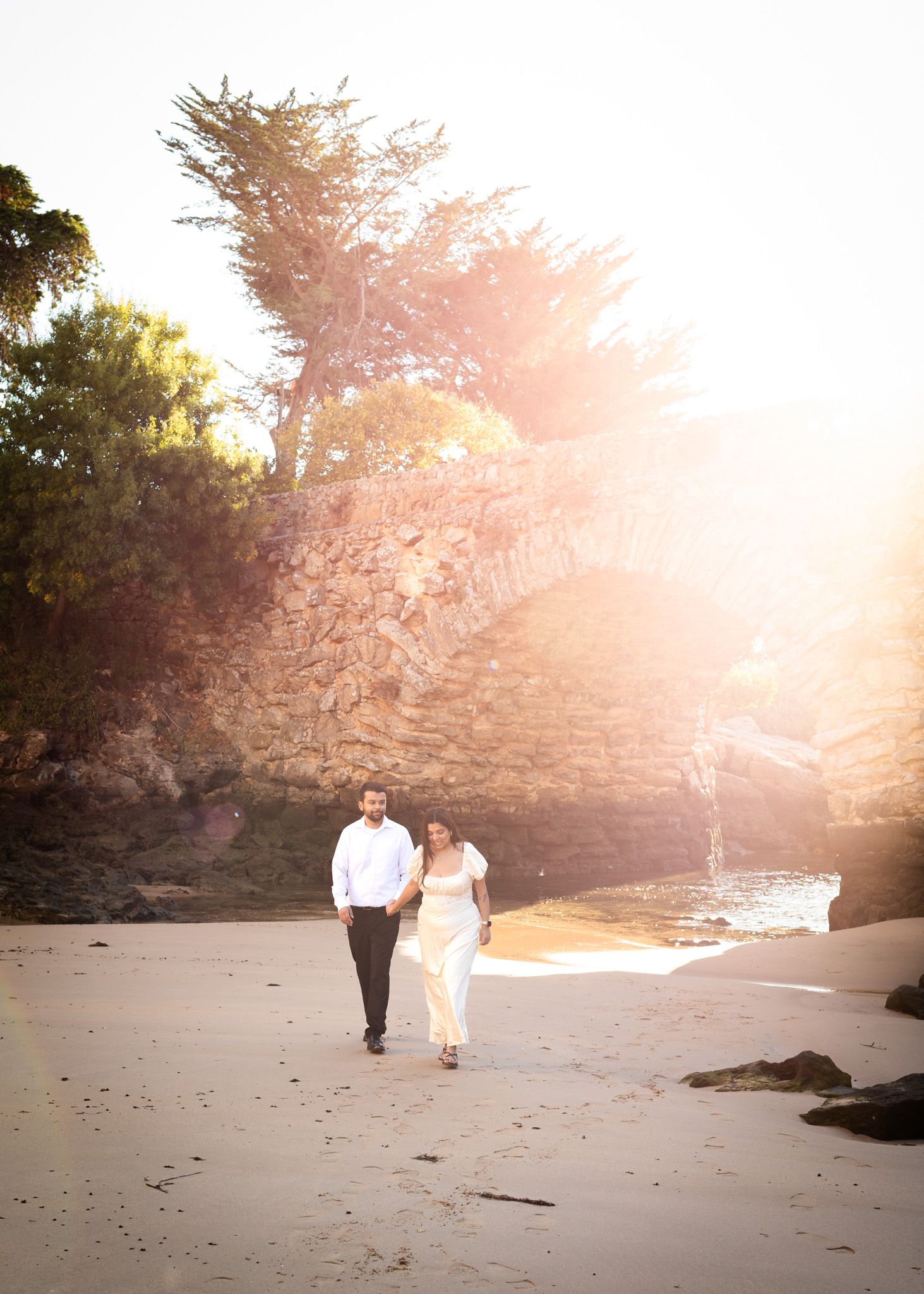 Couple walks hand in hand on sunlit beach with dramatic rocky cliffs and cypress trees.