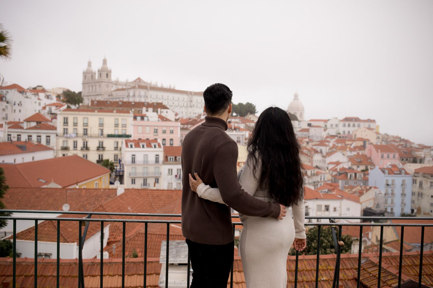 Couple embraces on balcony overlooking colorful Lisbon cityscape with terracotta rooftops.