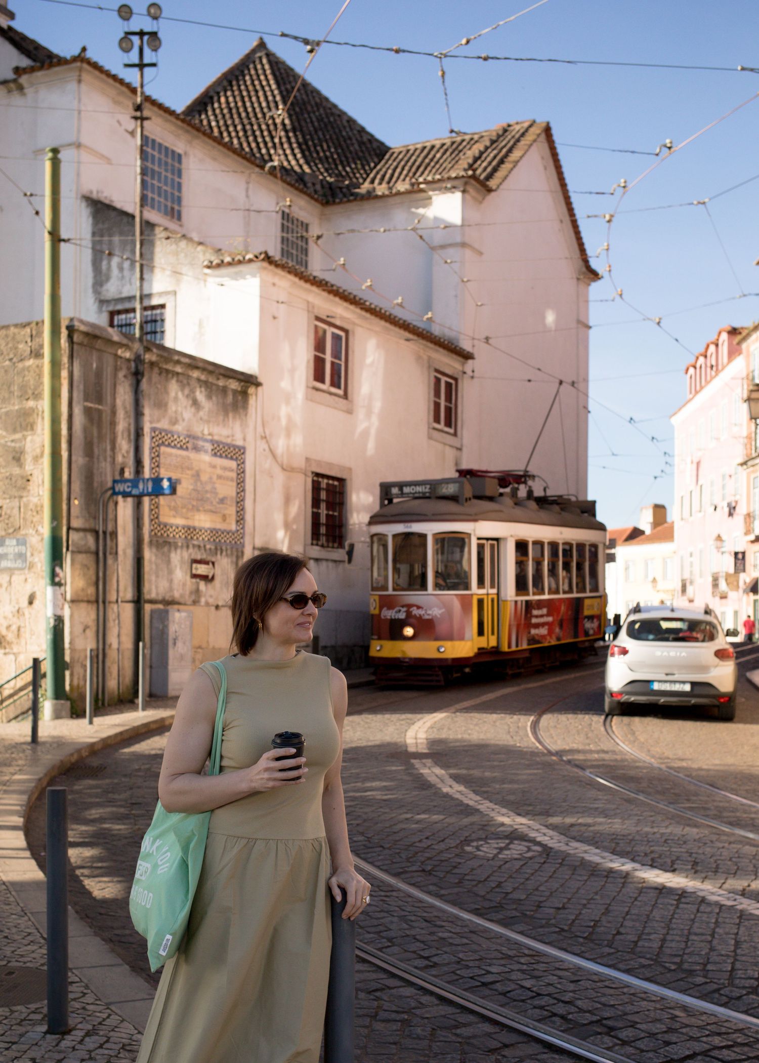 Woman in olive dress holding coffee walking Lisbon street with vintage yellow tram behind.