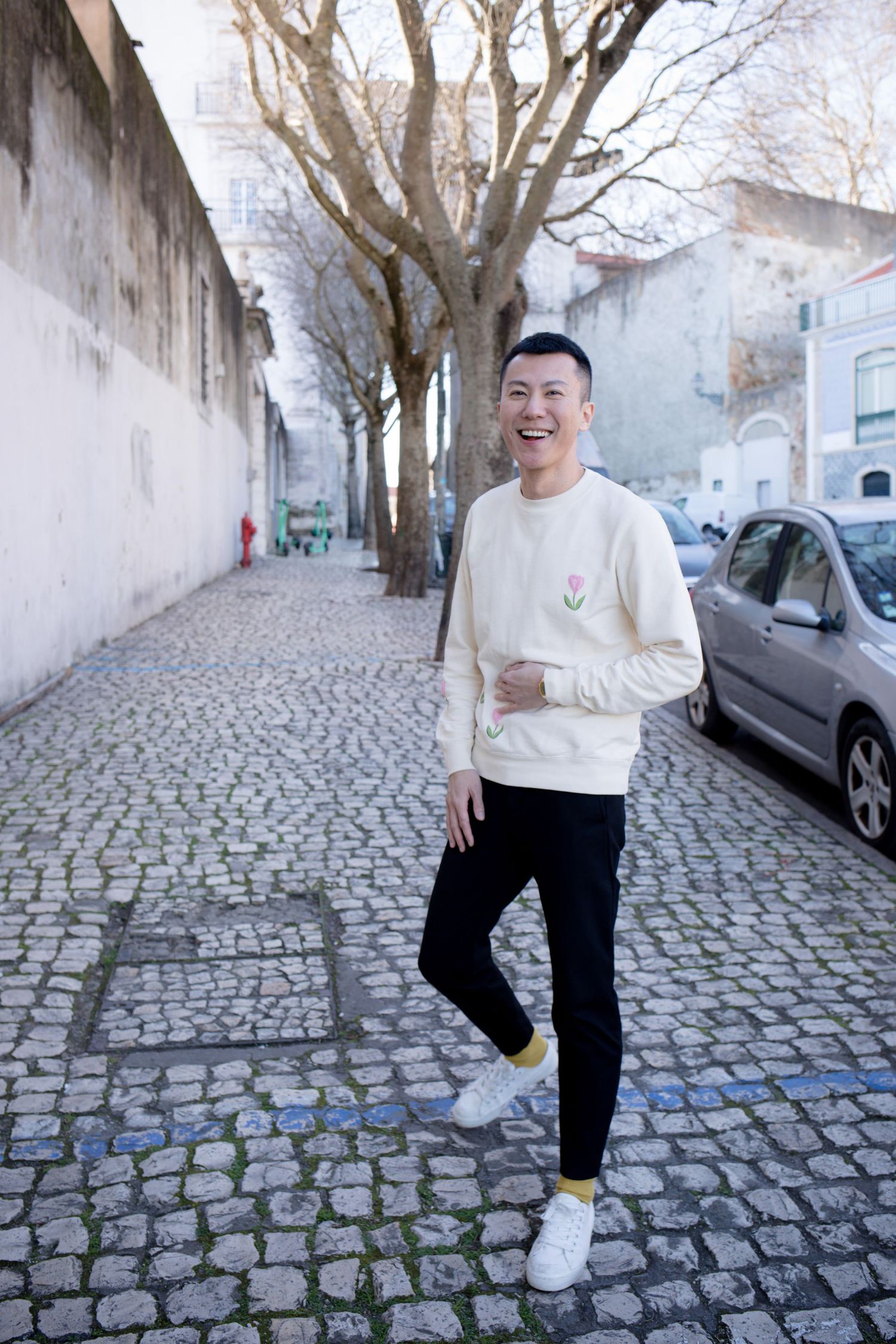 Smiling Asian man in cream sweatshirt standing on cobblestone street with bare trees.