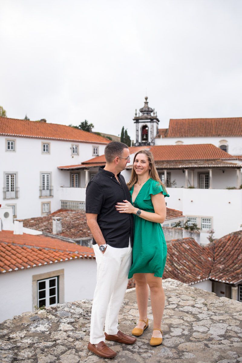 Couple embracing on a stone terrace overlooking red-roofed white buildings in a historic European town.