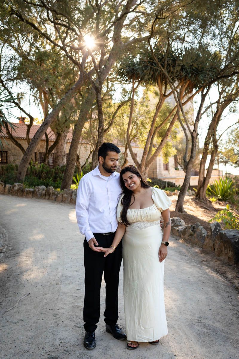 Couple posing on a sunlit path surrounded by trees, man in white shirt and woman in cream dress holding hands.