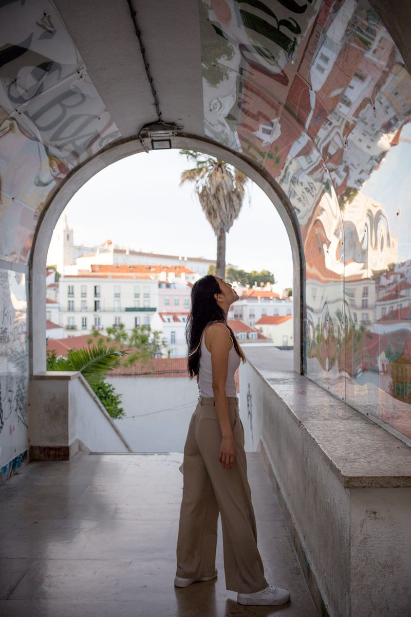 Woman in beige wide-leg pants stands under graffiti arch overlooking Lisbon cityscape with palm tree.