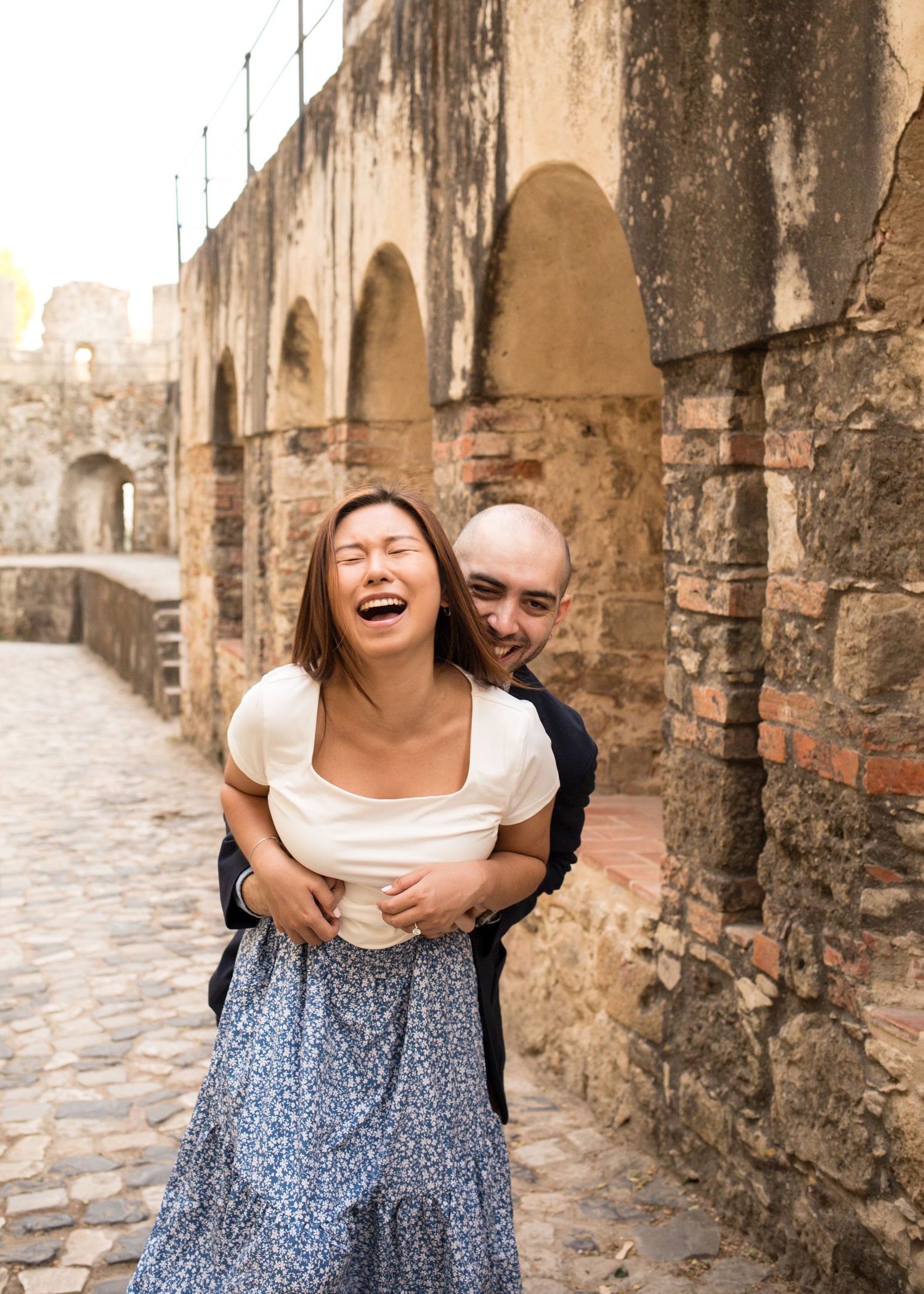 A laughing couple embraces near ancient stone arches on a cobblestone street, sharing a joyful moment together.