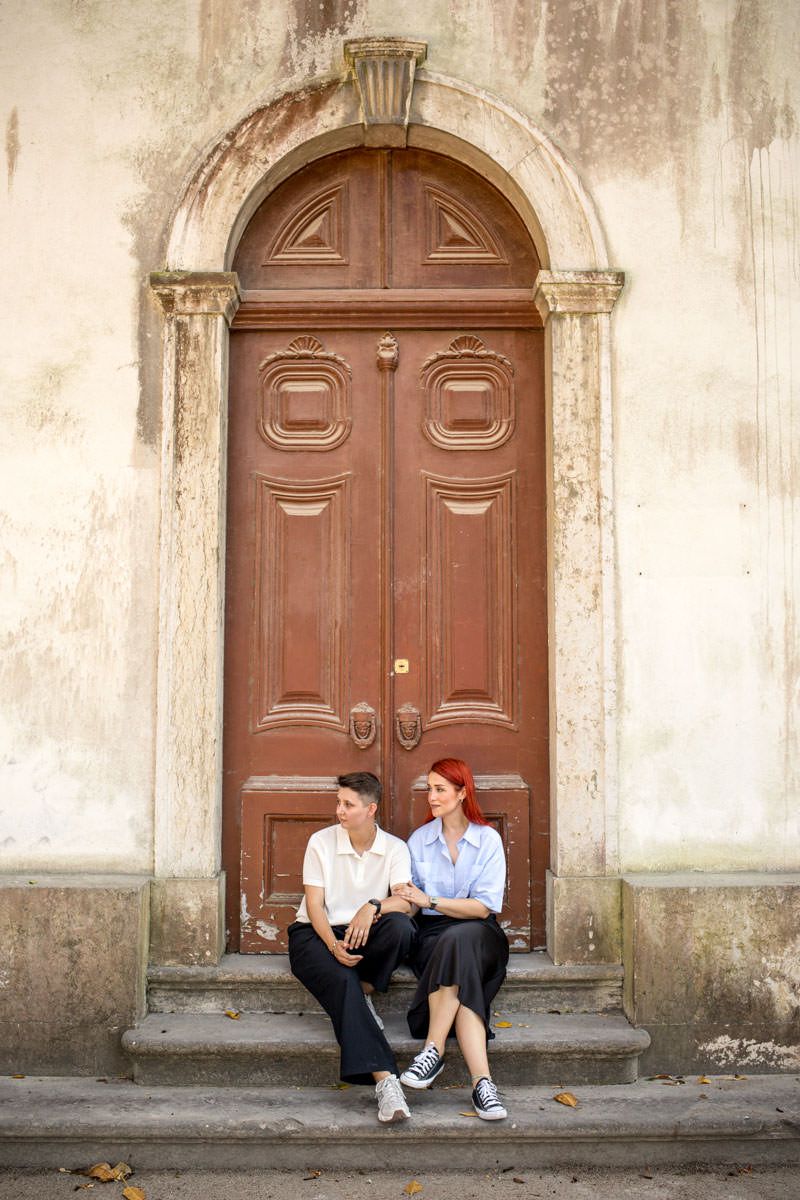 Two women sitting on steps in front of a vintage arched wooden door on an old building facade.