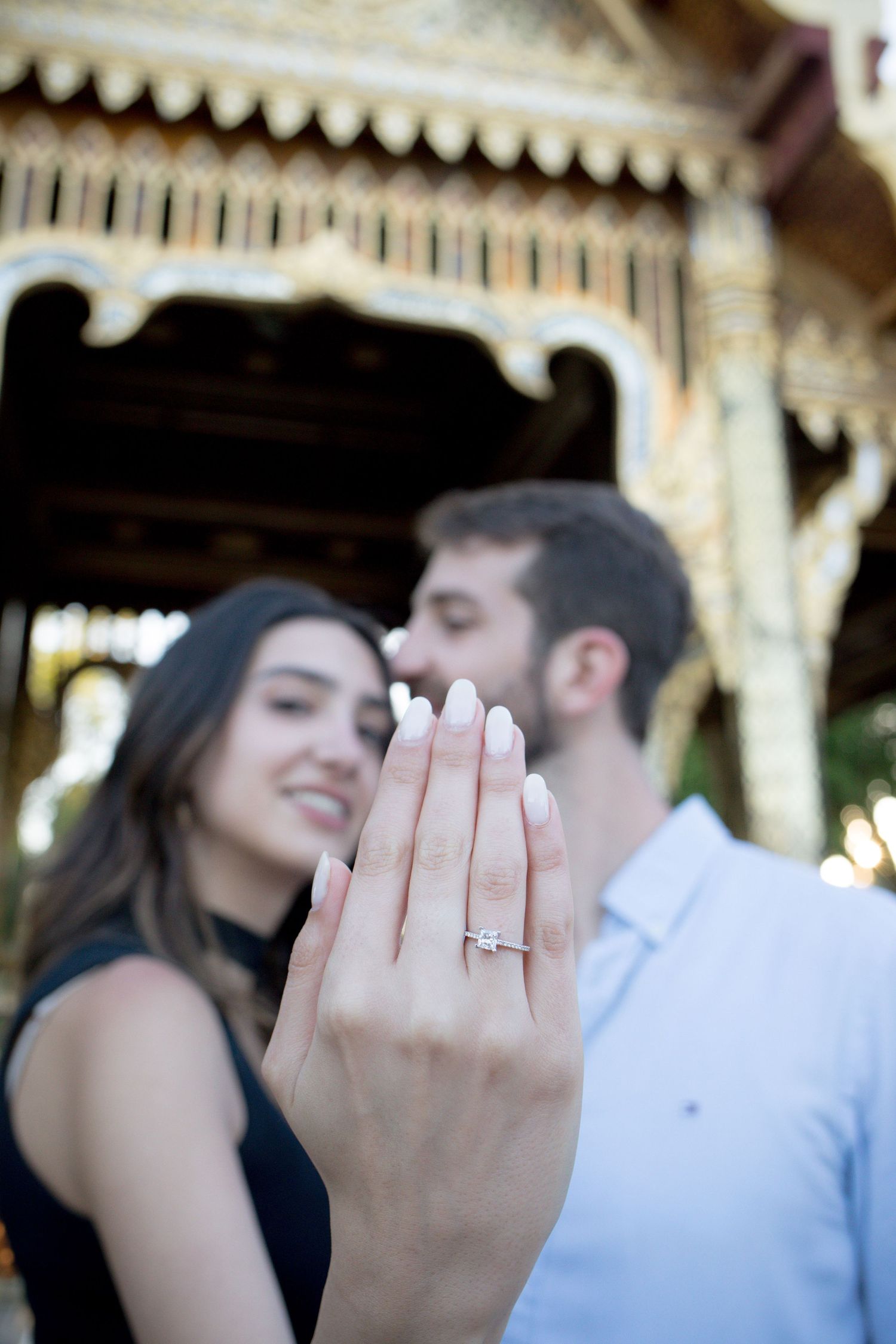 Couple showing off diamond engagement ring in front of ornate golden pavilion, woman smiling with manicured nails.