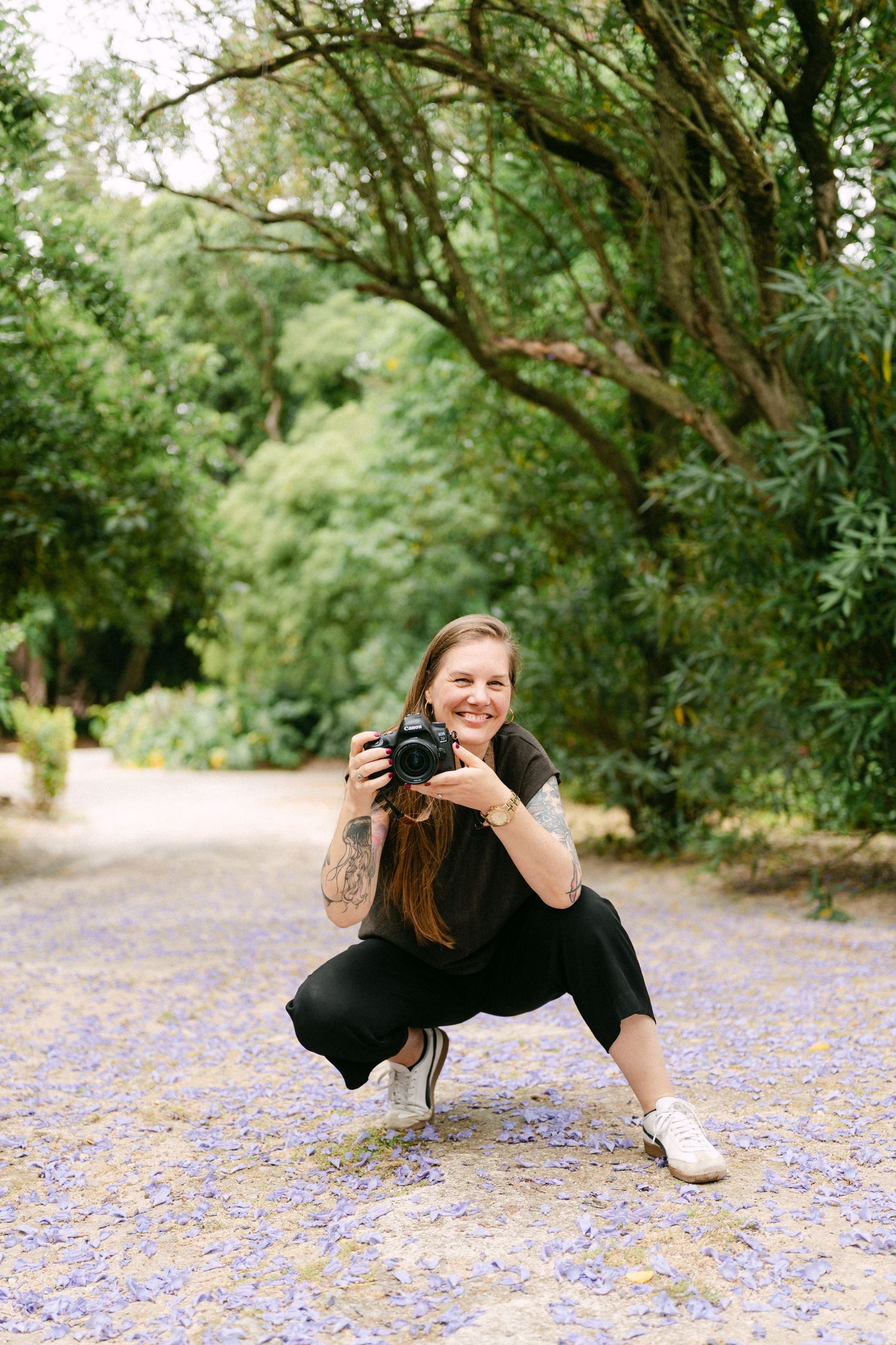 Smiling female Lisbon photographer crouching on purple flower-covered path in lush green park, holding DSLR camera with tattoos visible.