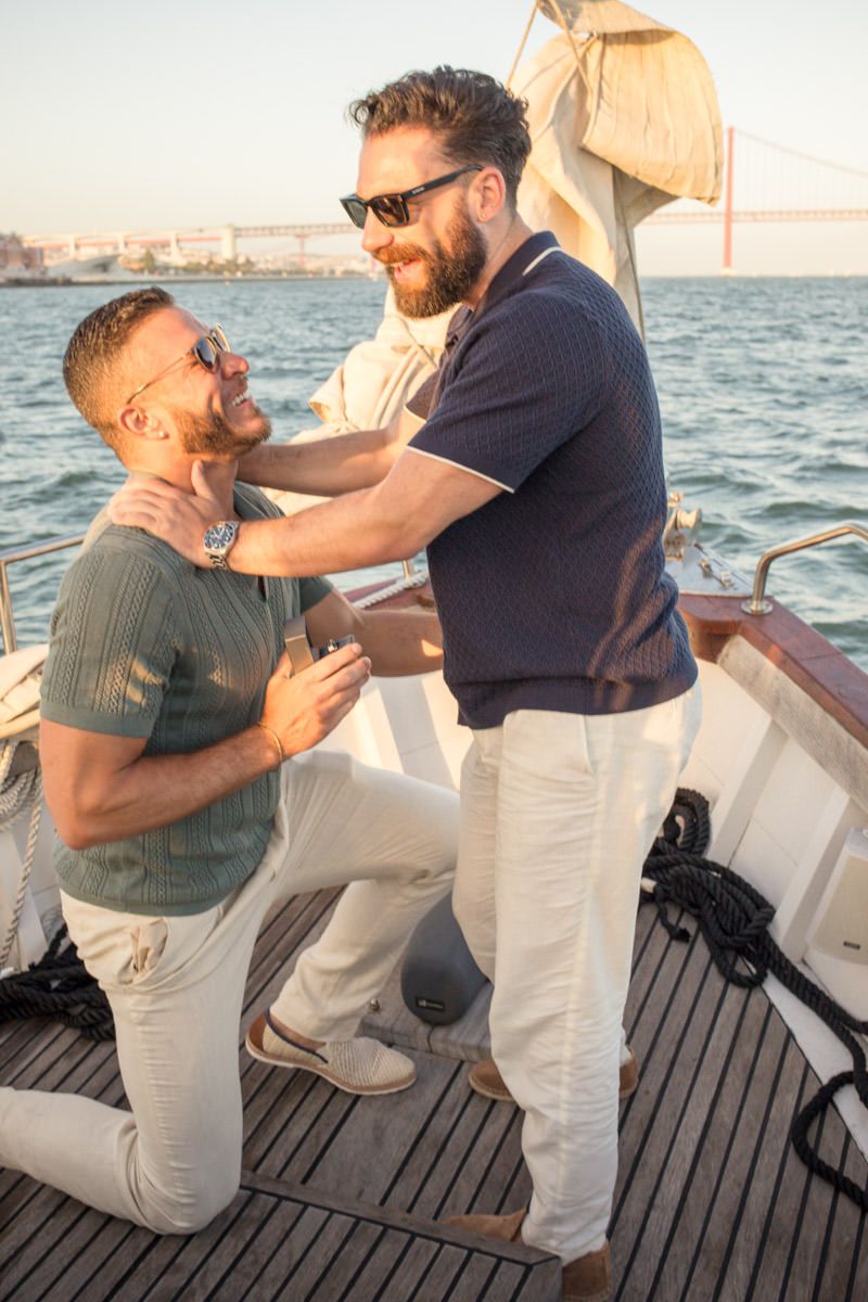 Two men on a sailboat at sunset, one kneeling as the other places hands on his shoulders.