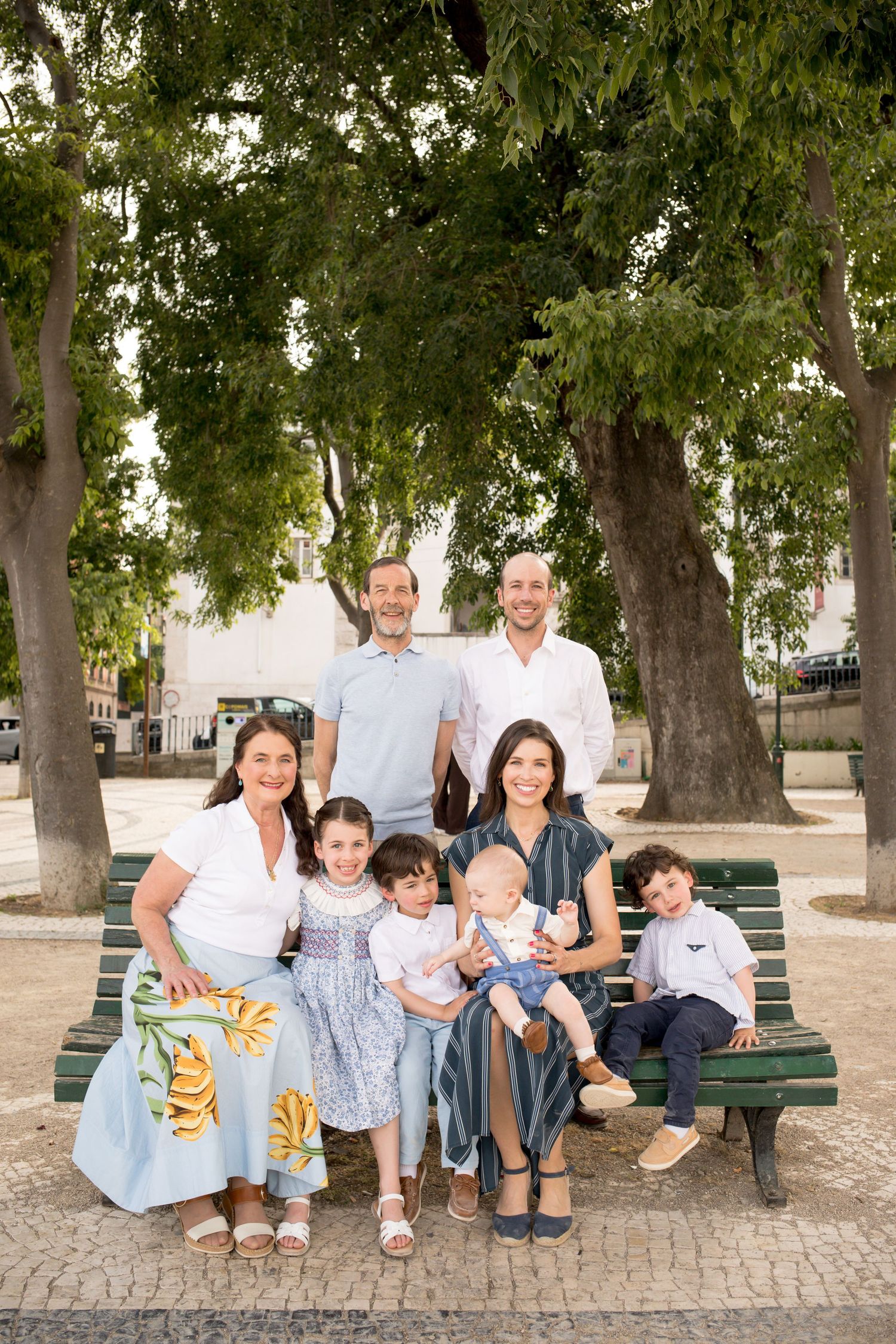 Extended family portrait on a green bench surrounded by lush trees in a Lisbon park.
