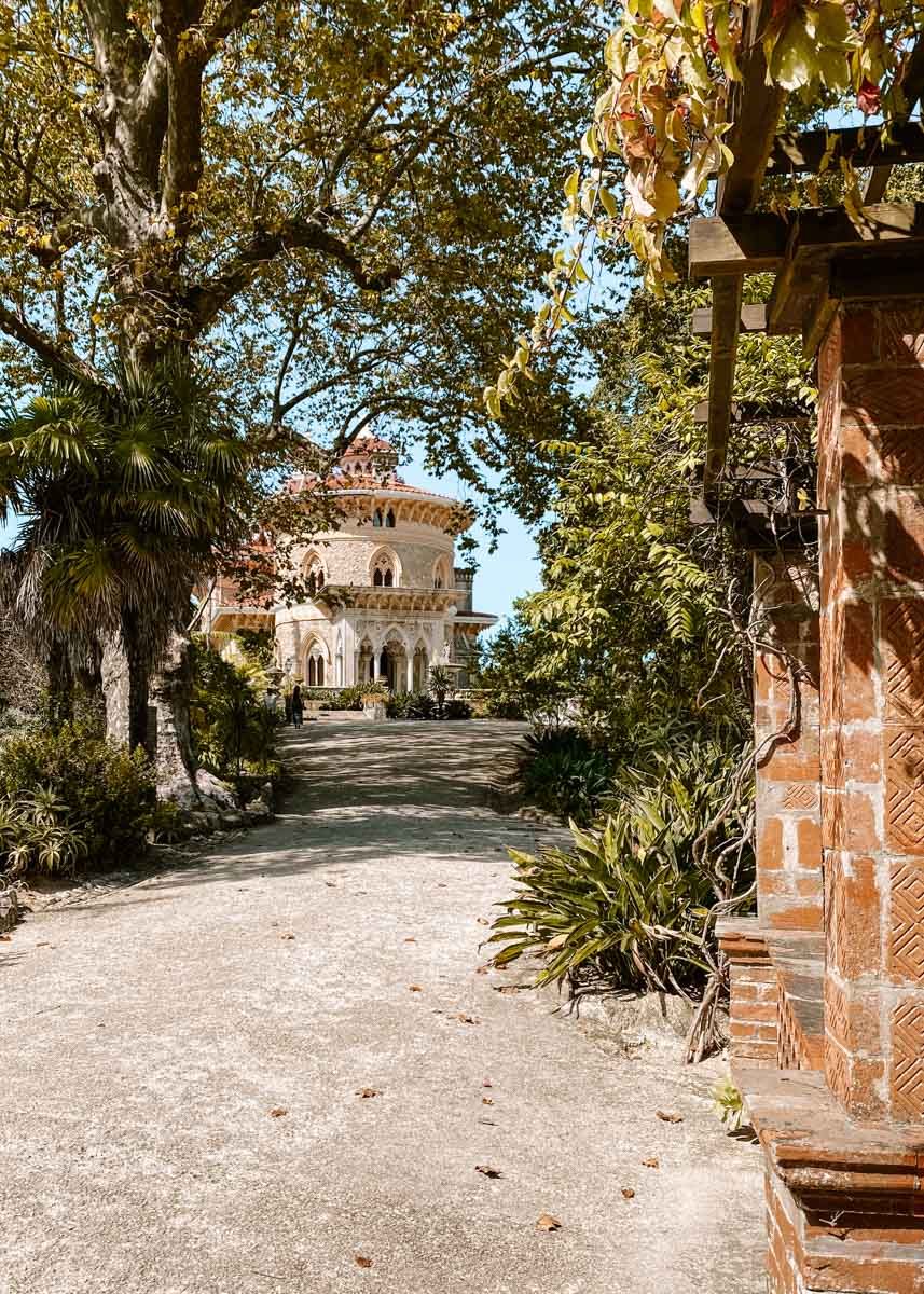 A tree-lined path leads to an ornate white palace surrounded by lush tropical gardens in Sintra, Portugal.