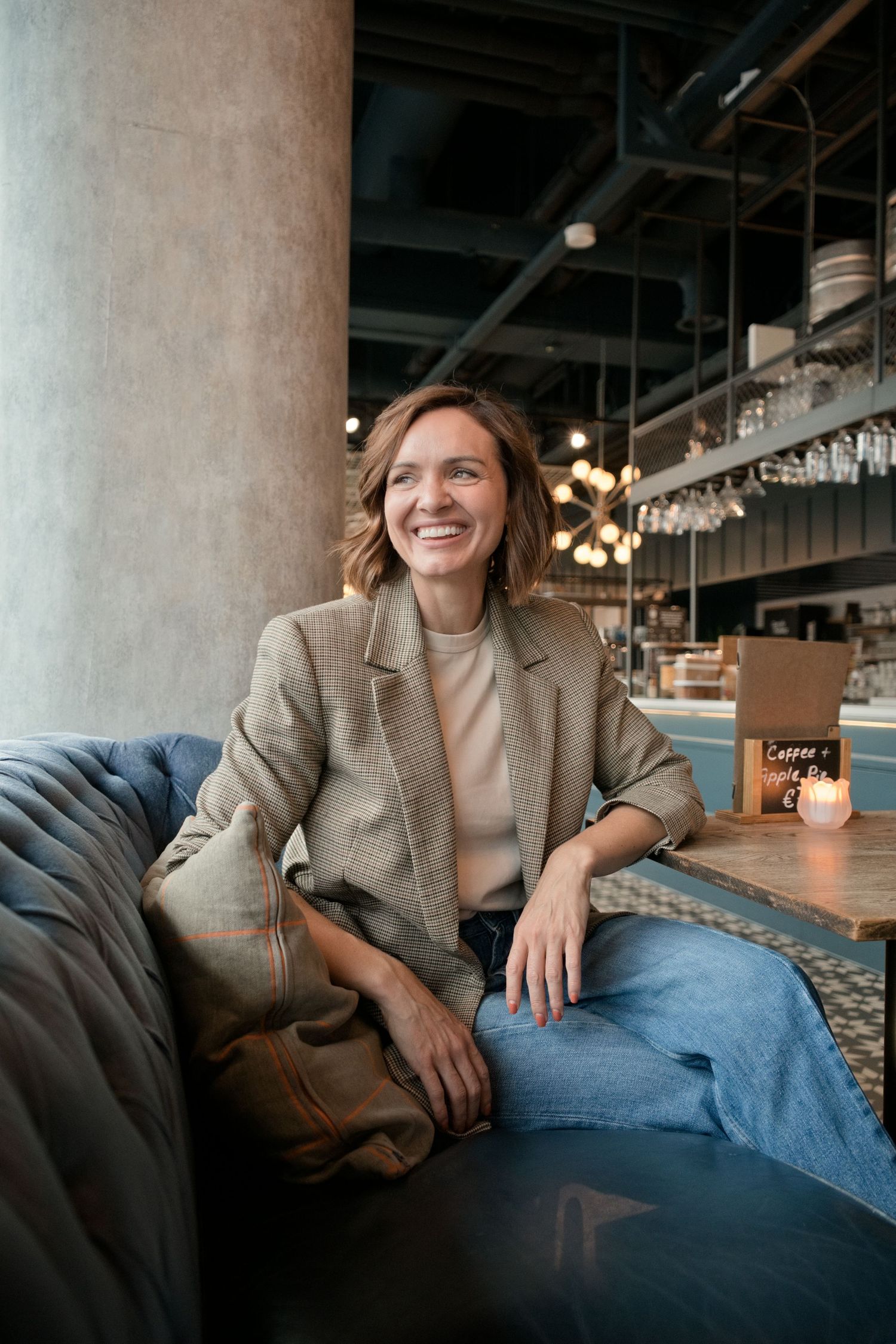 Smiling woman in a tweed blazer and jeans relaxes on a tufted leather sofa in a stylish industrial café with warm string lights.