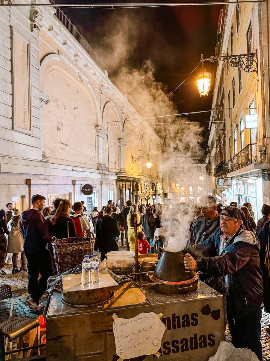 Street vendor roasting chestnuts at a steaming cart on a bustling European cobblestone street at night.