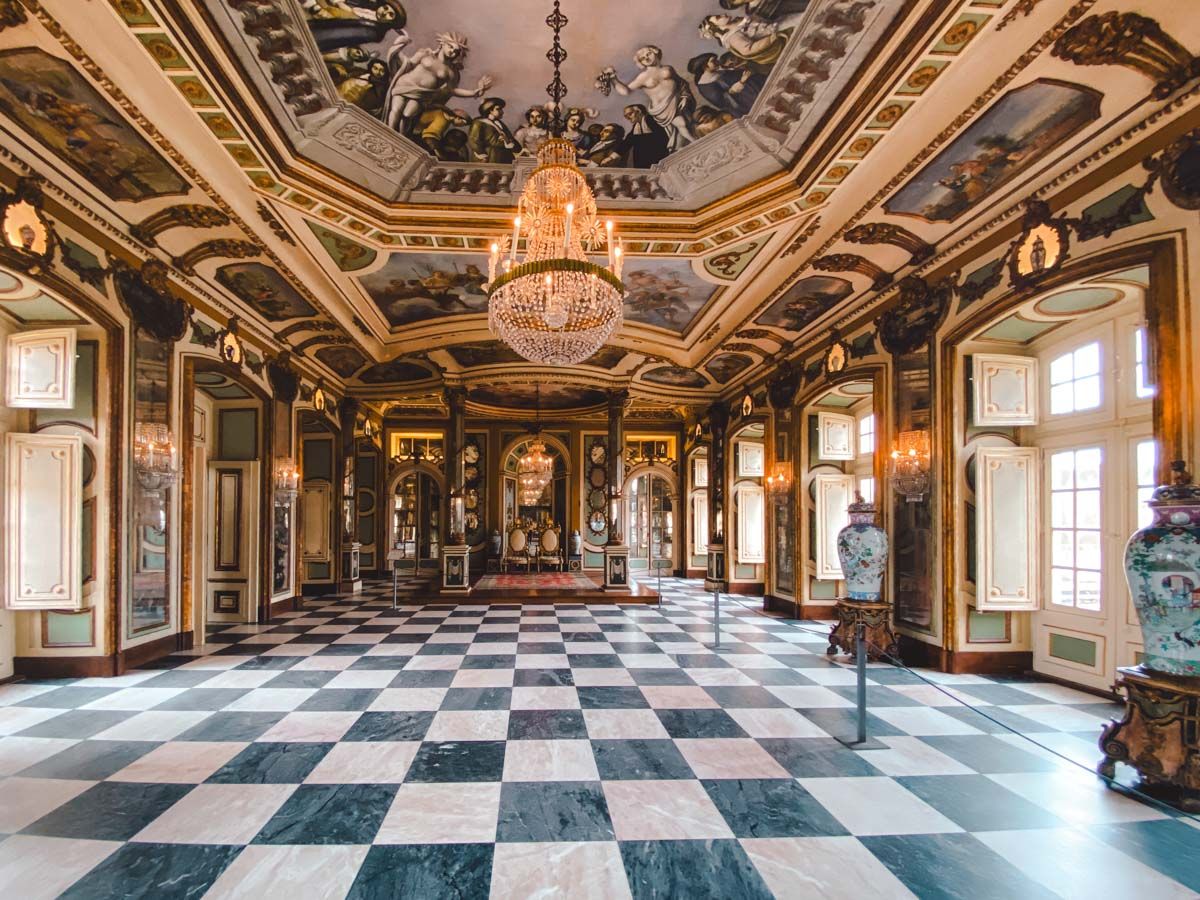 Ornate palace ballroom with checkered marble floors, gilded baroque ceiling murals, crystal chandelier, and elegant mirrors.