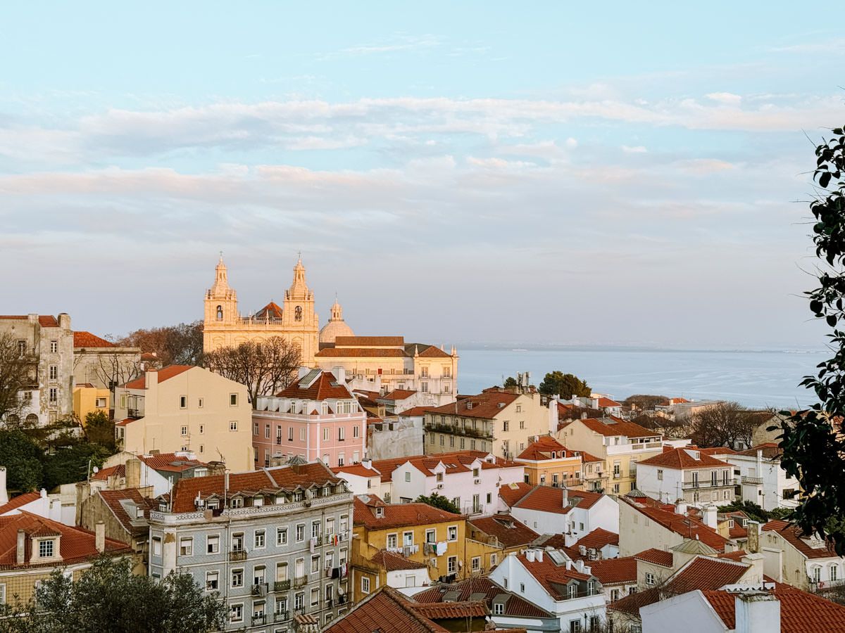 Panoramic view of Lisbon cityscape with terracotta rooftops, historic church, and ocean horizon at golden hour.