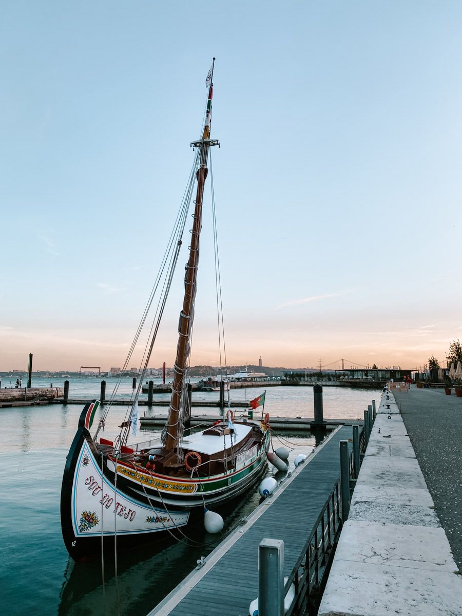 A vintage wooden sailboat docked at a calm marina pier during a serene sunset with soft pastel skies.