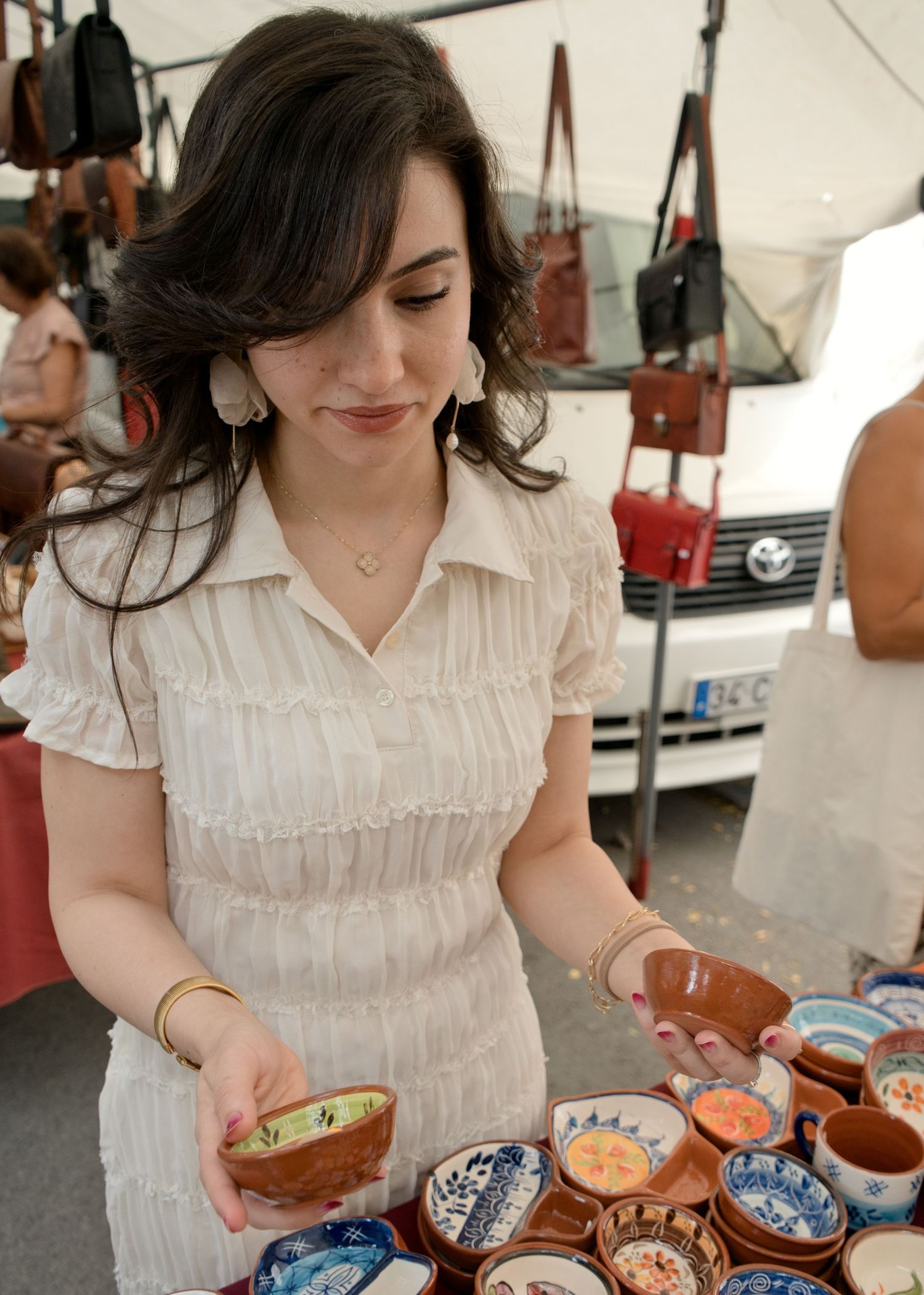 Woman in a white ruffled dress holds a handmade ceramic bowl while browsing colorful painted pottery at an outdoor market stall.